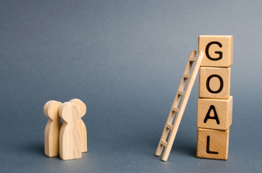 three wooden people are standing near a tower of cubes with word Goal that has a ladder leaning against the word Goa