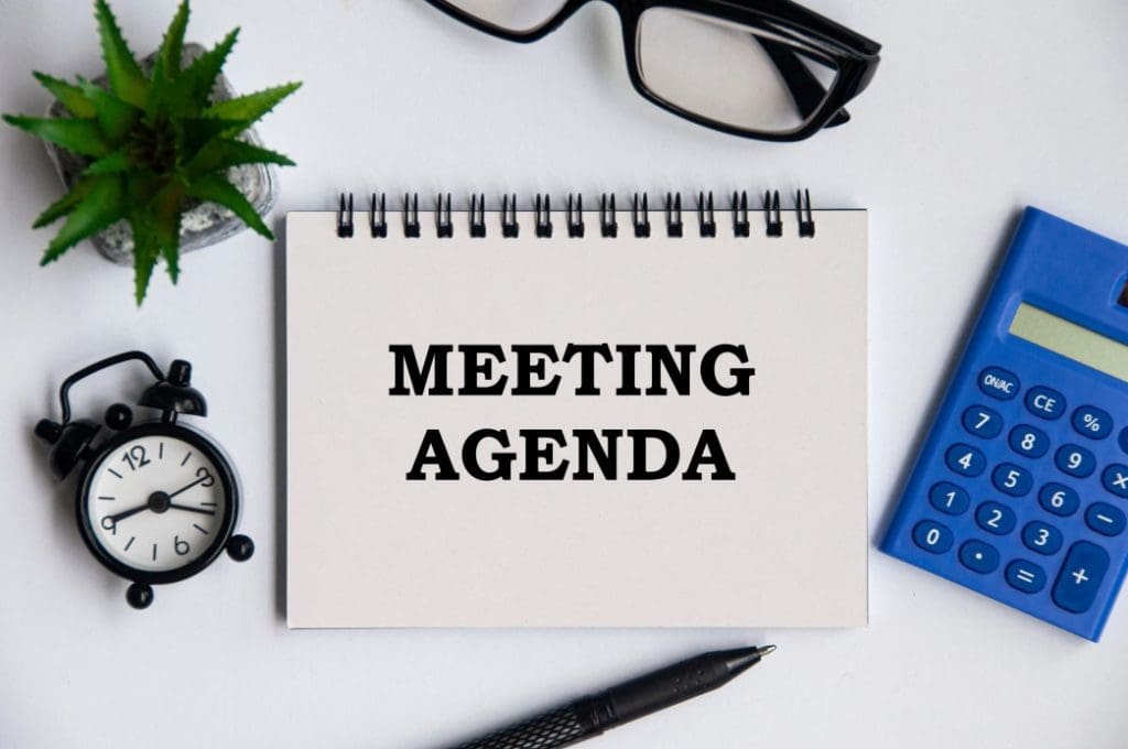 A Notebook with the words meeting agenda along with an alarm clock, calculator, small house plant and eyeglasses on a white table