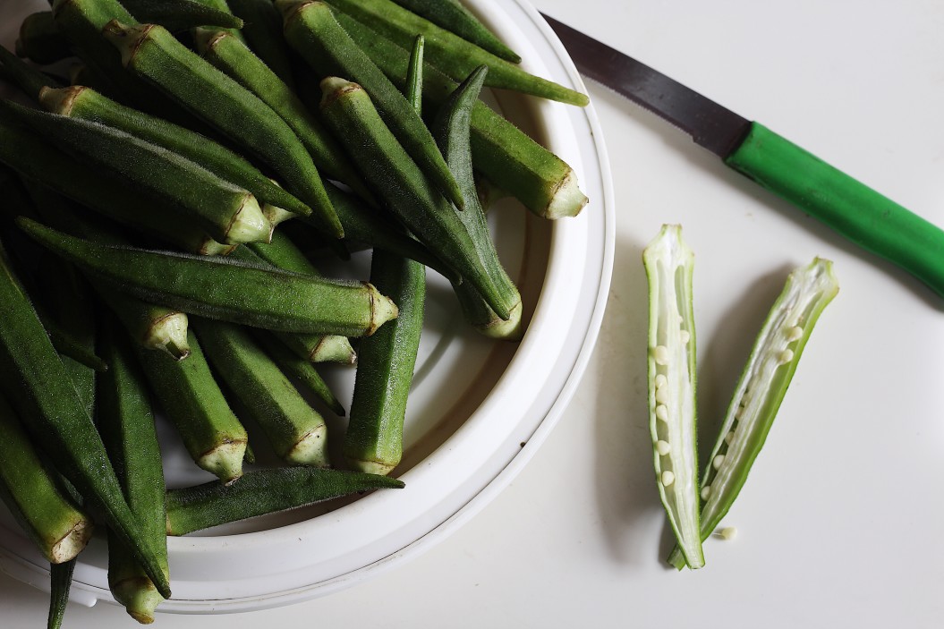 A bowl containing fresh okra and one okra on the table sliced in half and a knife next to it