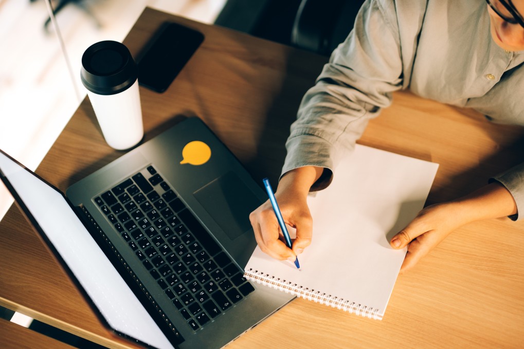 young woman studying online courses with a laptop writin gdown notes in a notebook and drinking coffee