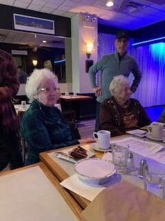 Two elderly women sit at a restaurant table with coffee and dessert, while a standing man looks on.