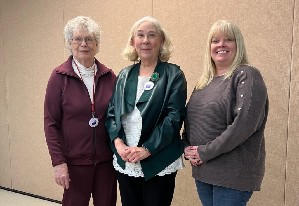 Three women stand indoors against a beige wall, facing forward. One has curly white hair and glasses, another has blonde hair and wears a green jacket, and the third has long blonde hair and a gray sweater.