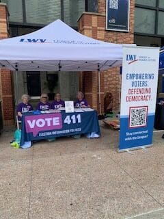 Four people sit at a table under a tent with "VOTE 411" and League of Women Voters banners, offering election information outside a brick building.